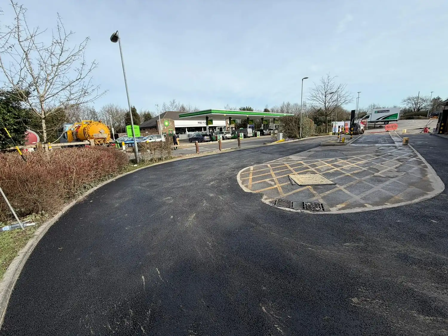 A newly paved gas station area with vehicles, trees, and road signage in a clear sky setting. Construction equipment is visible.